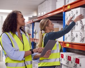 Female Team Leader With Digital Tablet In Warehouse Training Intern Standing By Shelves