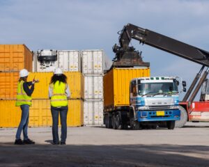 Two female workers in safety vests and hard hats oversee container loading operations at a shipping yard. A crane lifts a yellow container onto a truck, highlighting teamwork and global logistics.